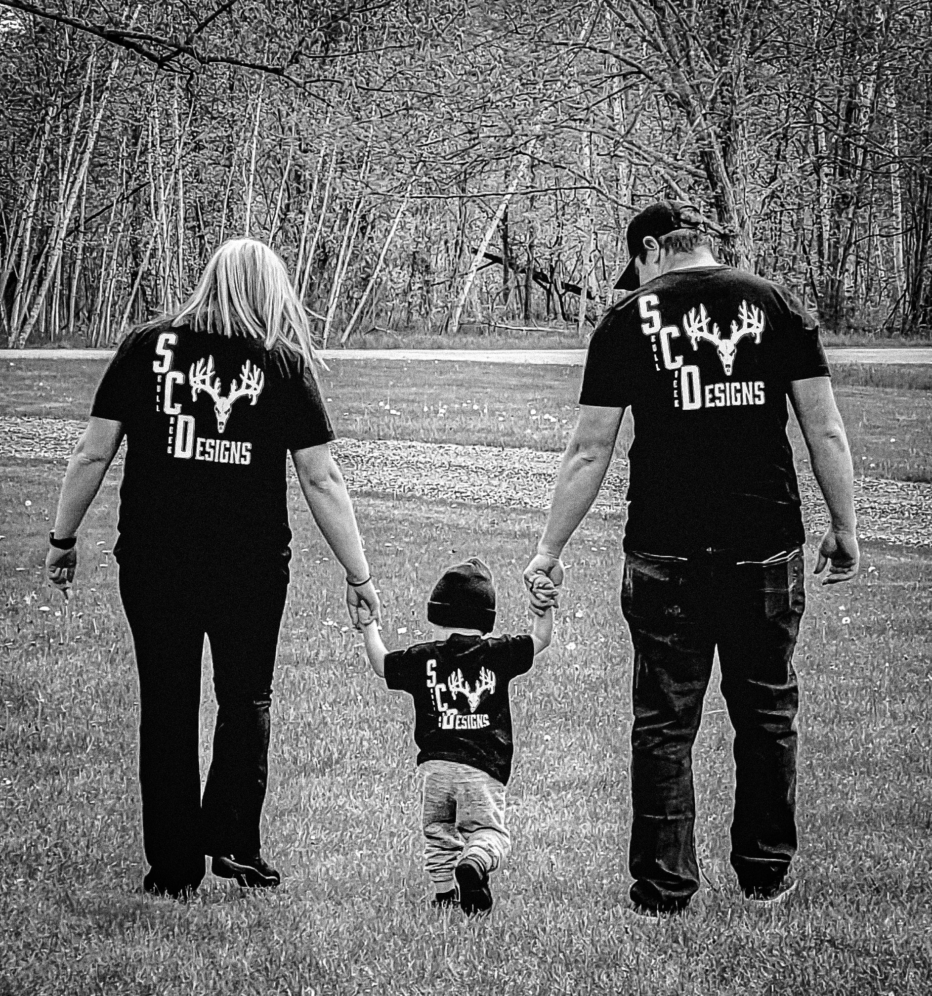 A black and white photo of two adults holding hands with a child, all wearing matching Skull Creek Designs shirts, walking away in a grassy area, symbolizing family values and unity.