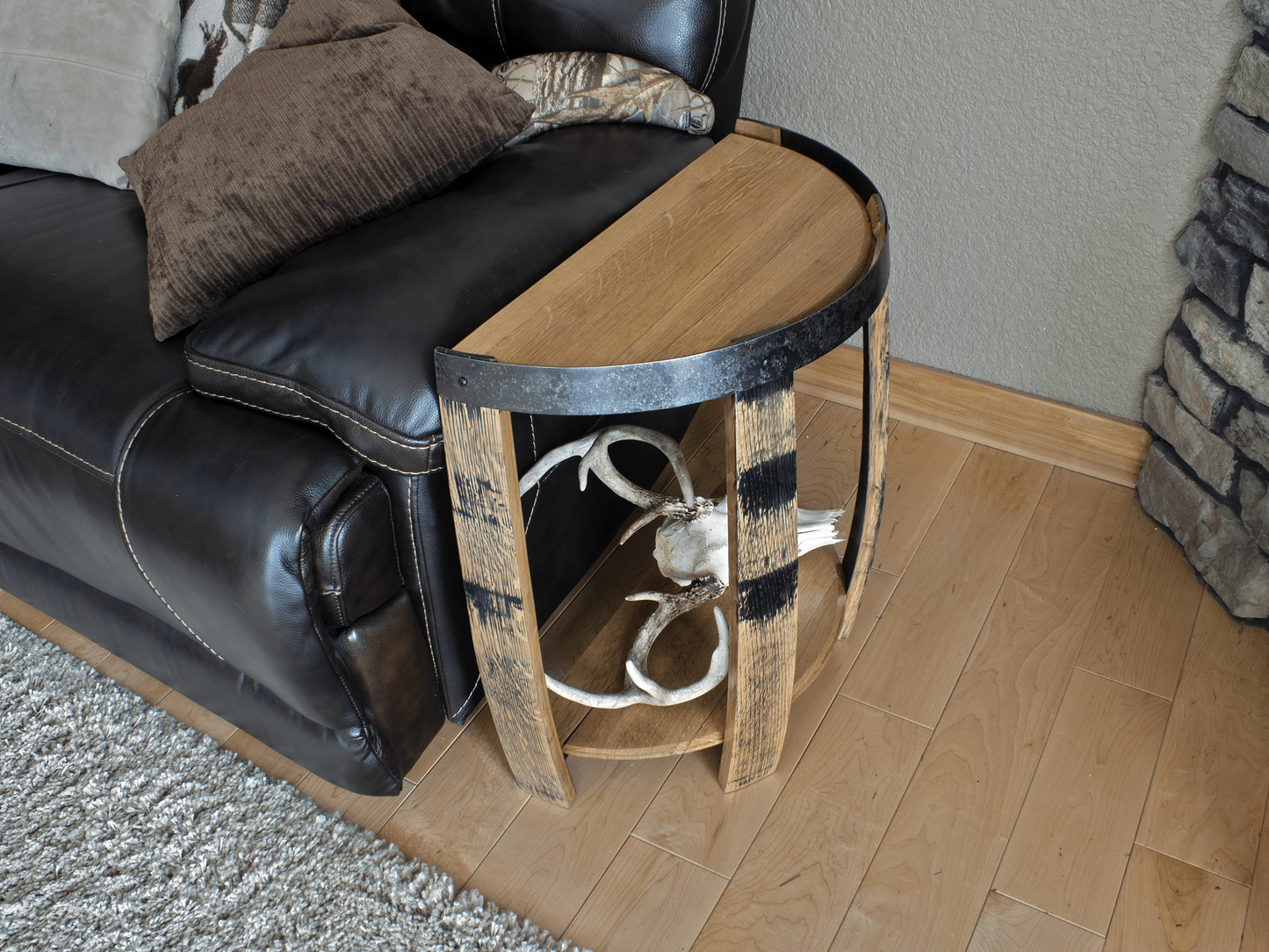 Overhead angled view of a rustic half whiskey barrel end table, showcasing the rich texture of the reclaimed barrel wood and contrasting metal straps against a leather couch and wooden flooring.