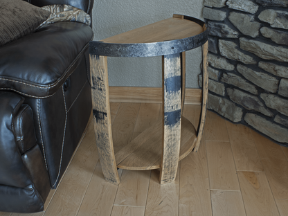 Angled view of a reclaimed whiskey barrel end table, showcasing the juxtaposition of the rustic, charred wood staves against the polished metal band, positioned next to a leather couch and against a textured stone wall for a stylish home aesthetic.