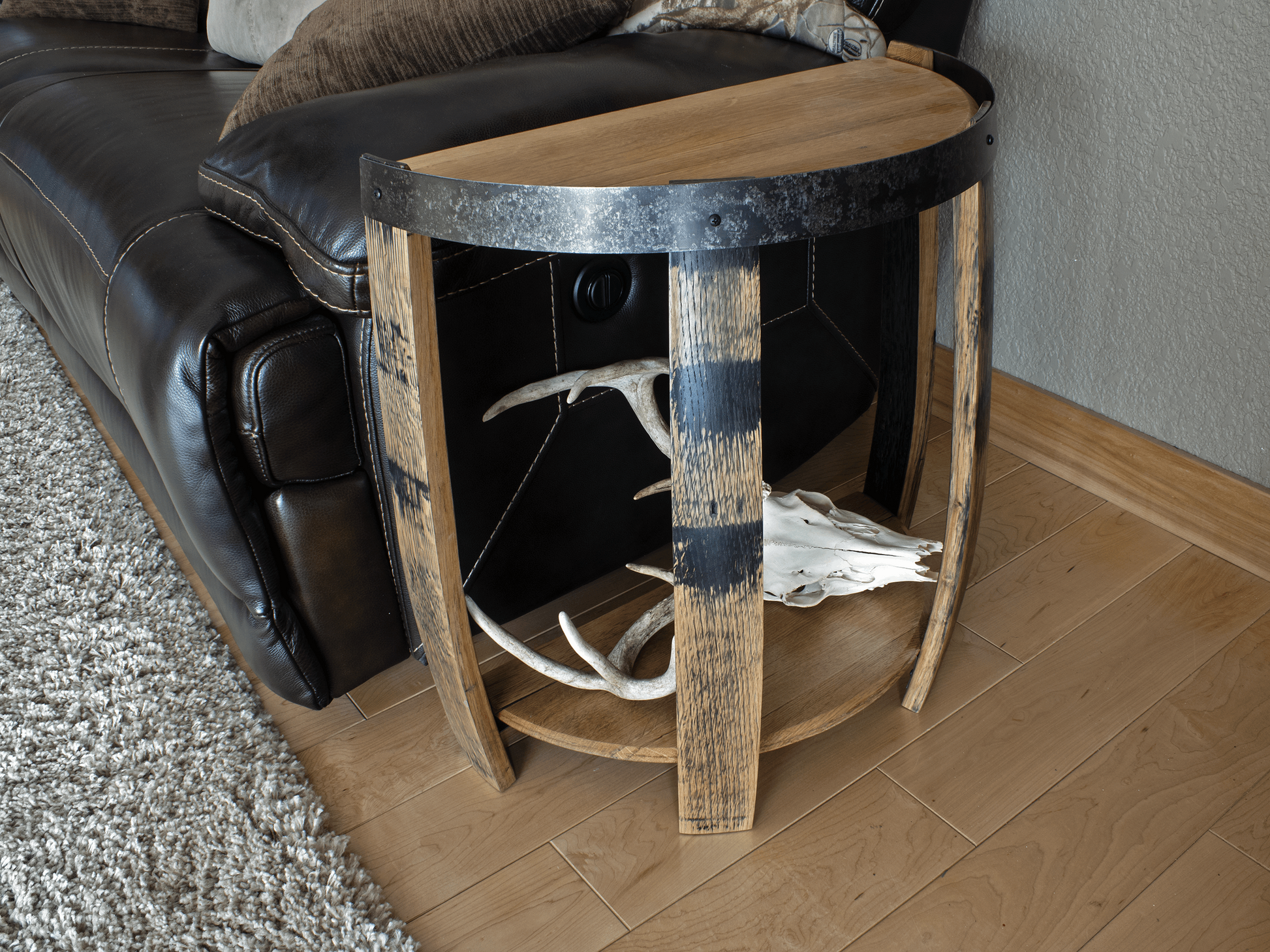 Side view of a handcrafted half whiskey barrel end table, displaying the unique charred wood finish and metal hoop detailing, alongside a leather sofa and soft rug, enhancing the rustic charm of the home decor.