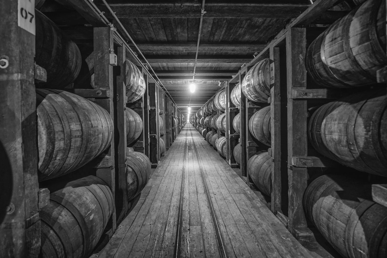 A black and white image of an aisle between rows of stacked whiskey barrels in a dimly lit warehouse, highlighting the rustic and authentic craftsmanship behind each product