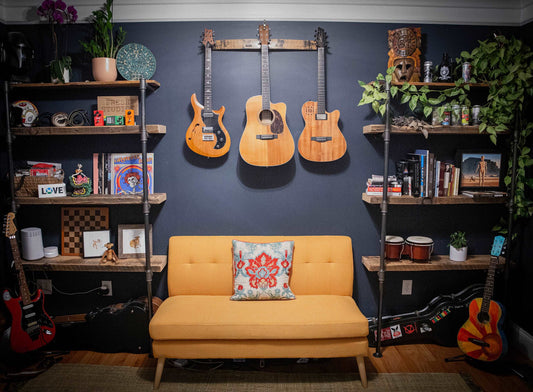 A 36-inch whiskey barrel stave guitar hanger displayed in a cozy music room, holding three acoustic guitars, with a vibrant mustard-yellow couch below and eclectic decor on surrounding shelves, combining functionality with rustic charm.